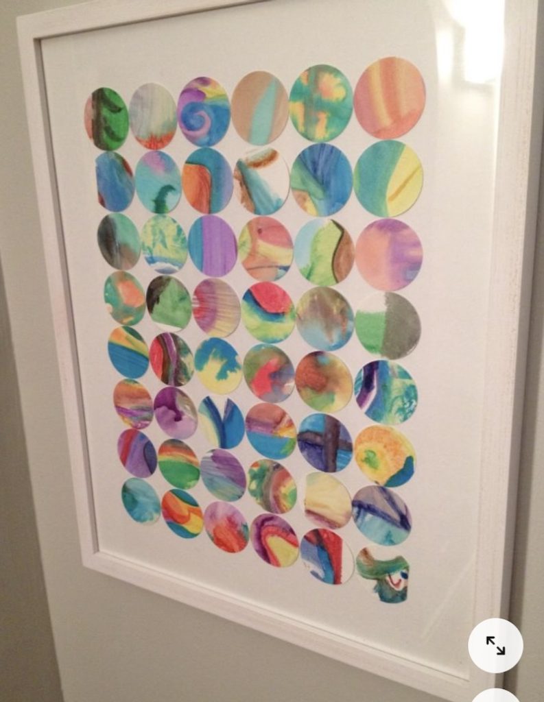 A framed artwork featuring a grid of colorful circles, each with a unique abstract pattern in watercolor-like shades of blue, green, orange, pink, and purple, hanging on a light gray wall.