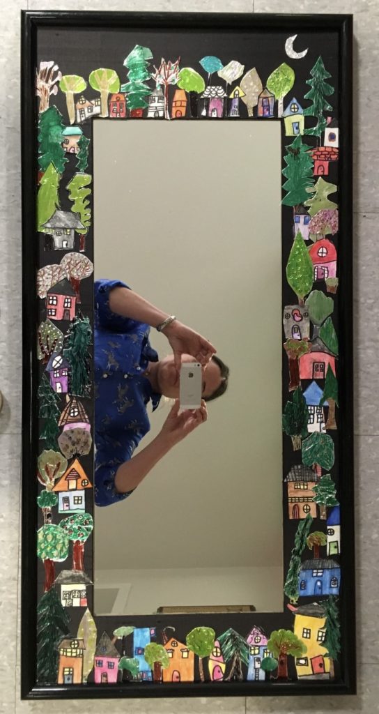 A person taking a photo in a mirror with colorful, hand-painted houses and trees decorating the mirror's border. The person is wearing a blue shirt and holding a smartphone.