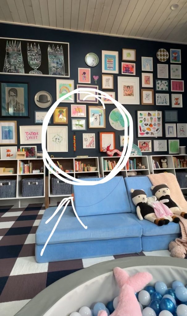 A colorful living room with a blue couch, stuffed toys, a ball pit, and a gallery wall filled with framed art and drawings. A large white circle and arrow highlight some childlike sketches on the wall.