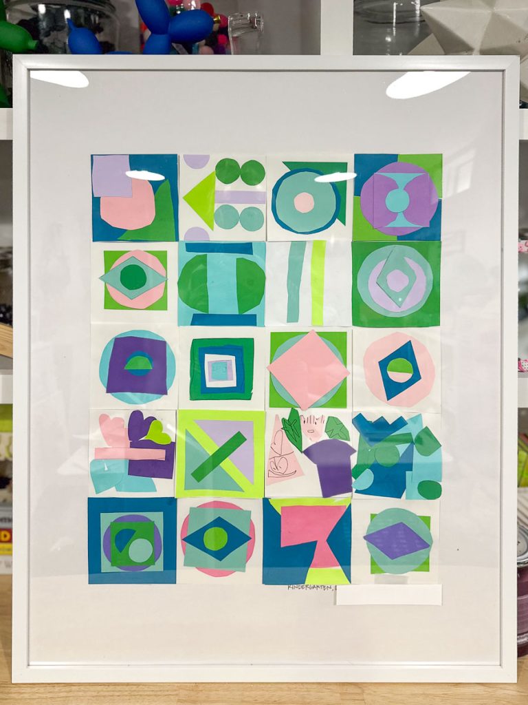 A framed abstract artwork made of colorful geometric shapes and patterns in various colors including green, blue, purple, and pink, arranged in a grid format. It rests against a shelf with various objects and decorations in the background.