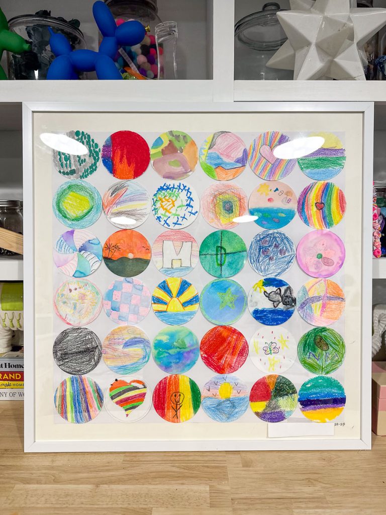 Framed artwork of colorful circular drawings by children. Each circle contains different patterns and colors, such as stripes, stars, and abstract designs, on a light background. Shelves with art supplies and decorations are in the background.