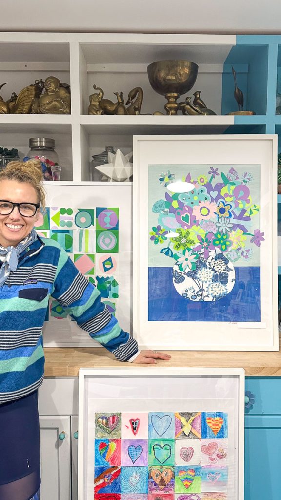 A person stands smiling by a table displaying colorful framed artworks. The artworks feature vibrant geometric shapes, hearts, and a floral design in a vase. The background shows shelves with decorative items, including bowls and small animal figures.