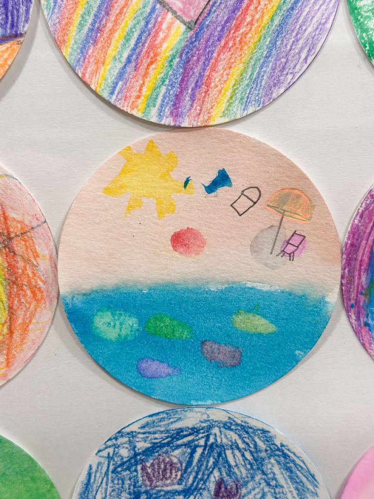 A circular drawing featuring a sunny beach scene with a bright sun, blue water, and colorful stepping stones. Two small figures, a beach umbrella, and a chair are also depicted. Surrounding circles show other colorful drawings.
