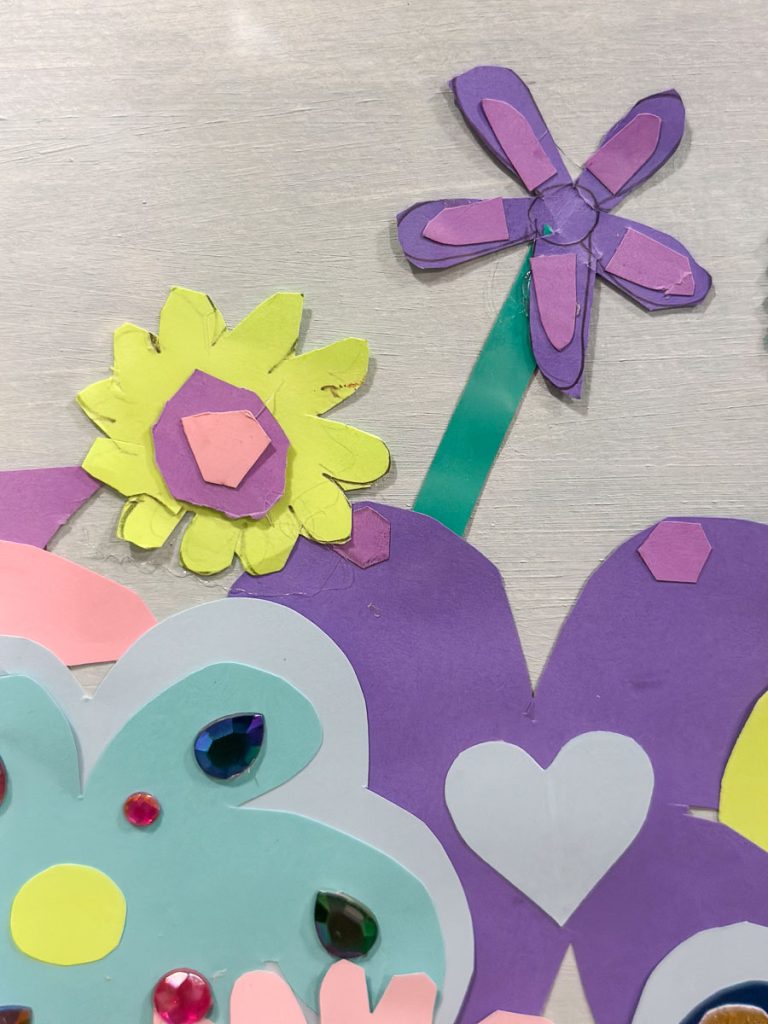 Colorful paper collage featuring purple, yellow, and pink flowers with a heart and gem decorations on a pastel backdrop. The mixed media arrangement includes various geometric shapes.