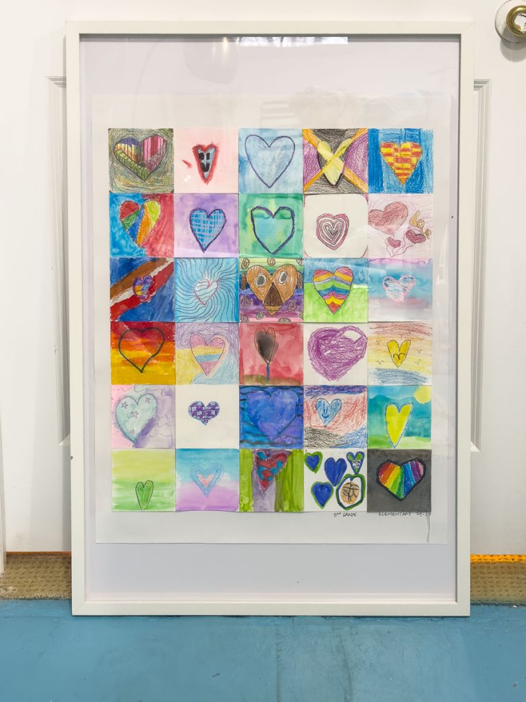 Framed artwork featuring a colorful grid of heart designs in various styles and colors, each unique in pattern and background, displayed against a white wall.