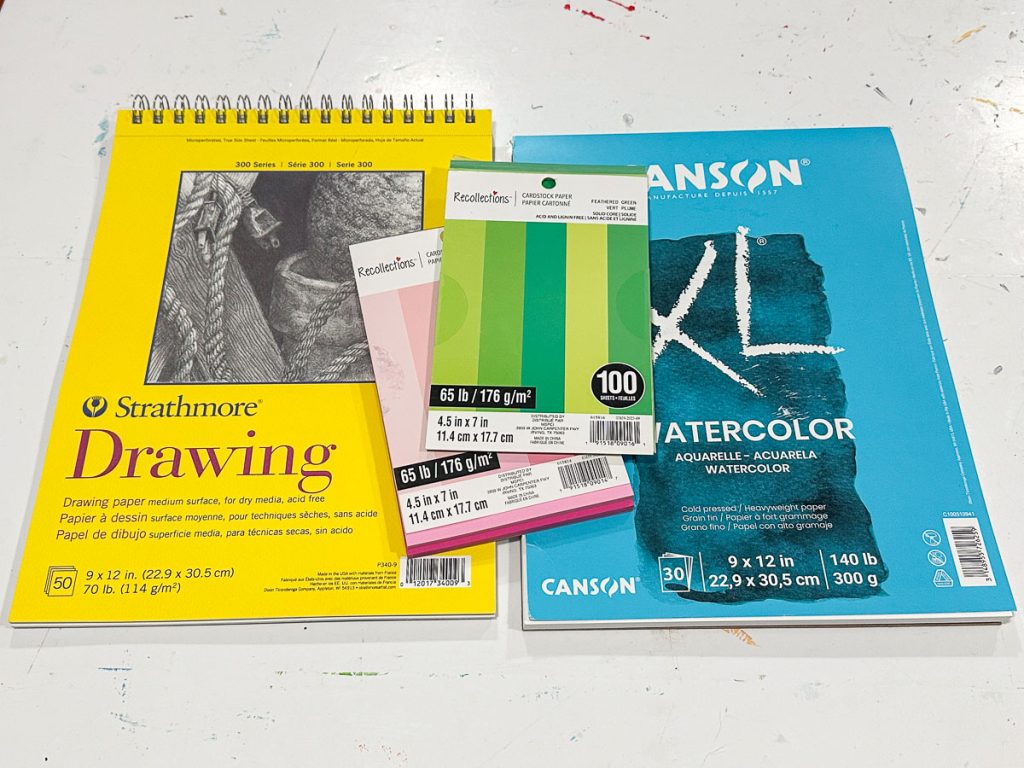 A collection of art supplies: a yellow Strathmore drawing pad, Canson watercolor paper pad, and two packs of colorful Reston Lloyd index cards. The items are on a white surface.