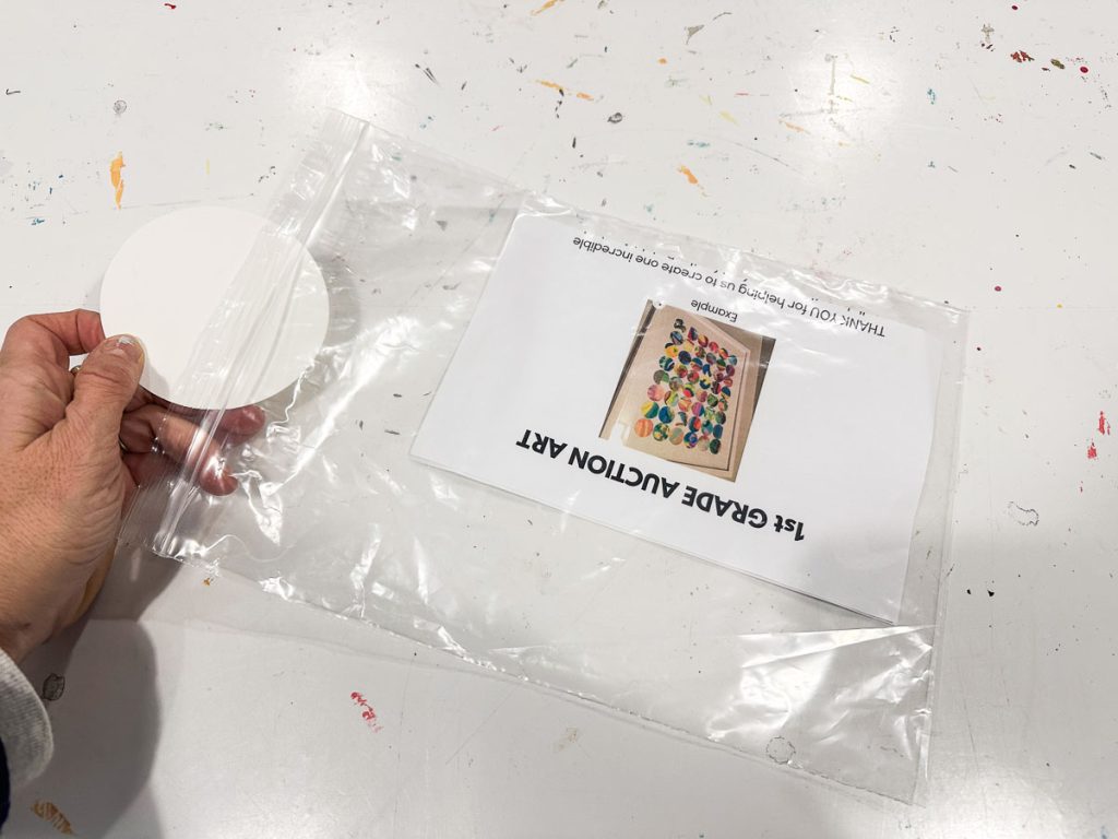 A hand holding an open plastic bag containing a round white disk and a sheet of paper with colorful artwork titled "1st Grade Auction Art" on it. The background surface is white with scattered paint marks.