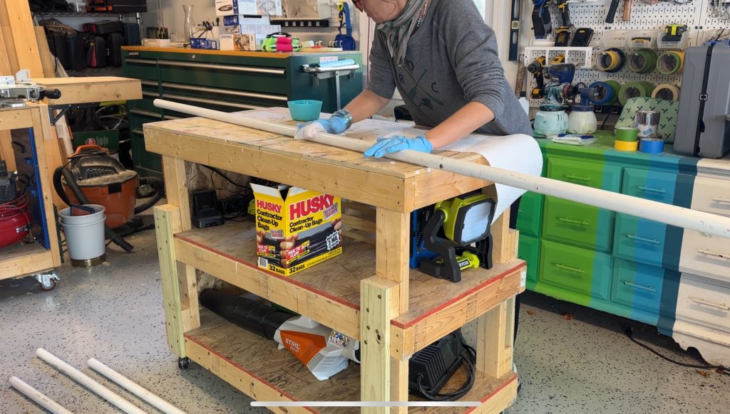 A person wearing gloves works on a long white PVC pipe at a wooden workbench in a workshop, surrounded by tools, boxes, and equipment.