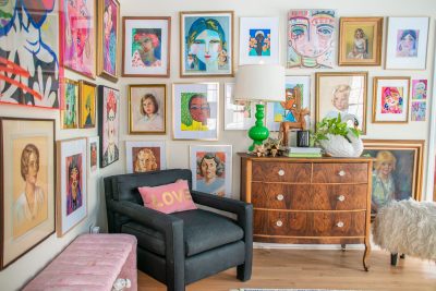 A cozy living room with a black armchair and a cushioned stool. The walls are adorned with vibrant portraits in various styles and frames. A wooden dresser holds a green lamp and decorative pieces, and there's a fluffy stool nearby.