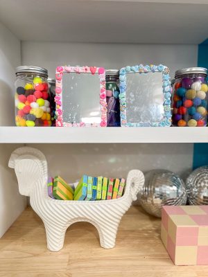 Shelves display jars with colorful pom-poms, two decorative mirrors with floral and cartoon frames, a white ridged llama-shaped container holding small notebooks or sticky notes, disco balls, and a pastel cube sculpture.