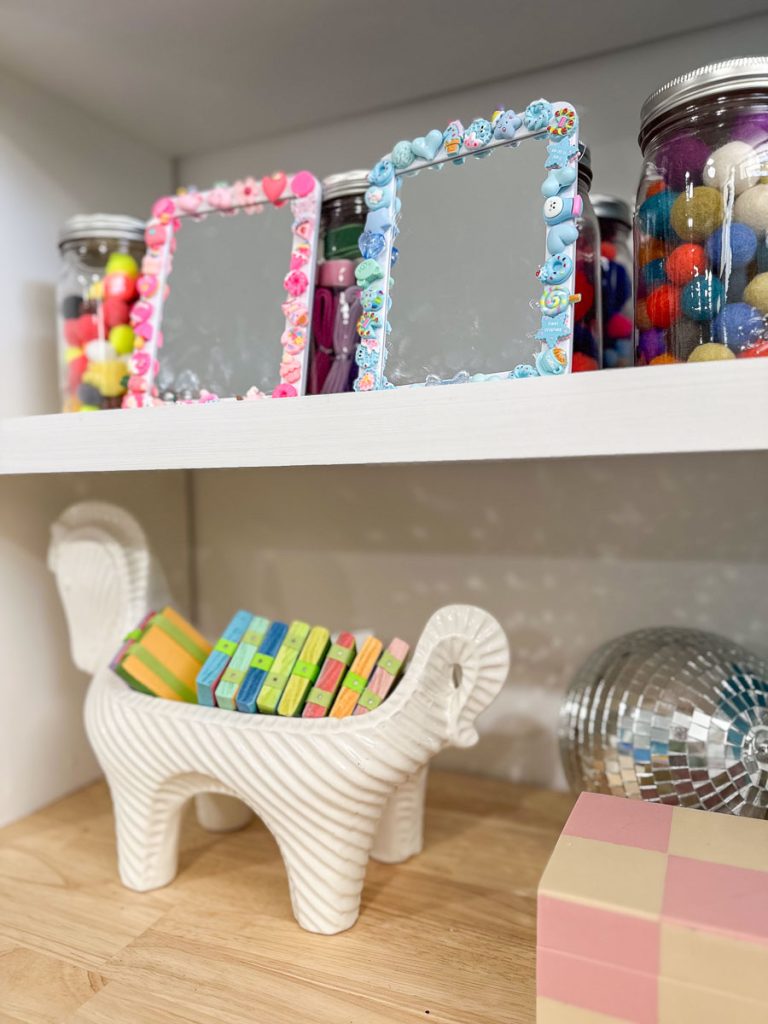 Decorative shelf with two colorful framed mirrors, jars of pom-poms, and a white ceramic animal figurine holding pastel-colored books. Nearby is a disco ball and a pastel checkered cube.