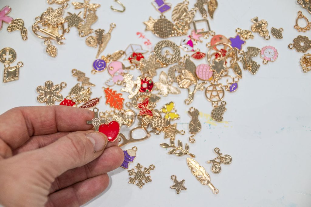 A hand holding a small golden charm with a red heart, amidst a scattered collection of assorted metallic charms on a white surface. The charms include various shapes like leaves, stars, and animals.