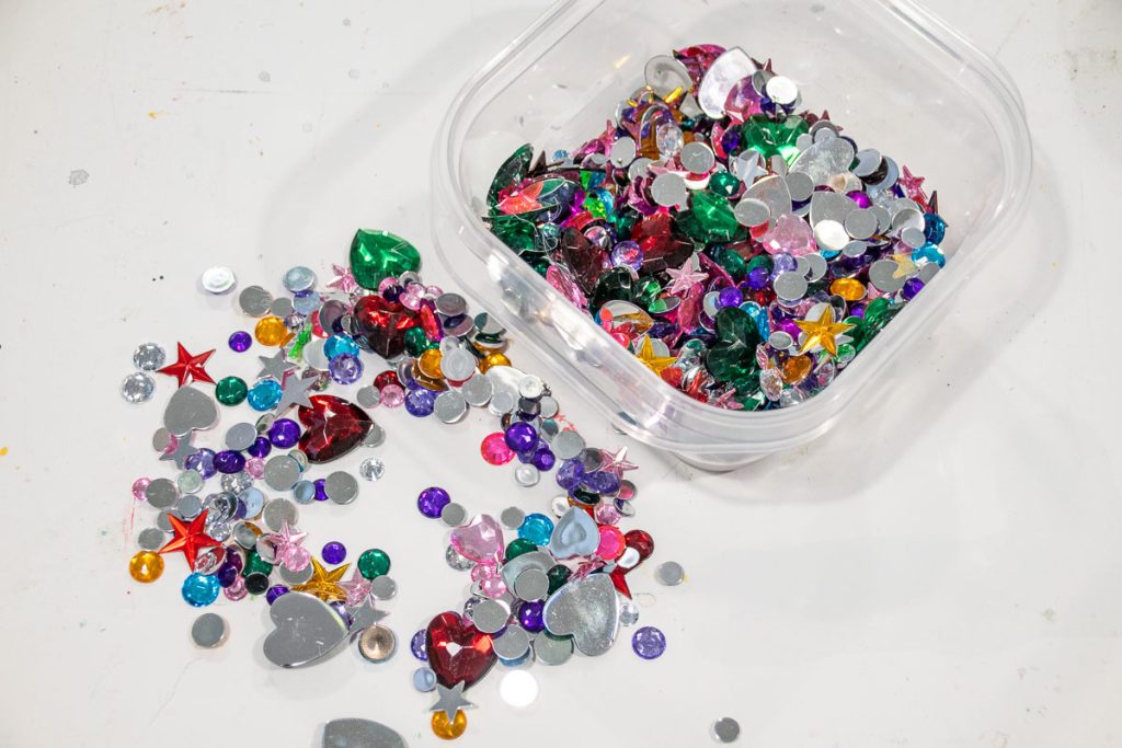 A clear container filled with colorful assorted craft gems, including stars, hearts, and round shapes, is on a white surface. Some gems have spilled out, creating a scattered, vibrant mix of colors and shapes.