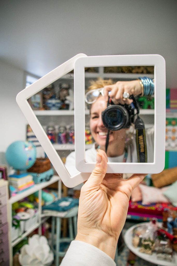 A person takes a photo of their reflection using a camera, capturing themselves smiling. They hold two mirrors, partially showing their face. The room is colorful, with shelves of figurines, a globe, and various items in the background.