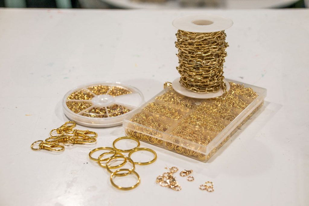 A spool of gold chain wire sits on a table, surrounded by a container with various gold clasps and rings. Some loose rings and clasps are scattered around. The setup is for jewelry making.