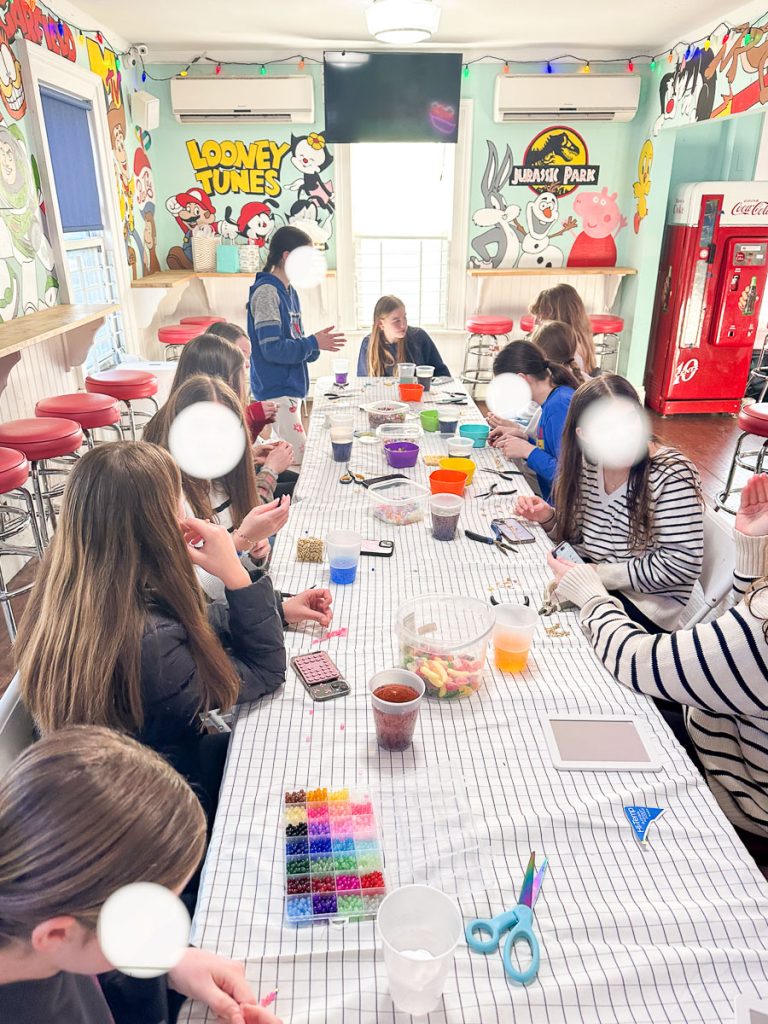 A group of people, mostly children, are gathered around a long table covered with a checkered cloth, engaging in a craft activity with beads and containers. The room is brightly decorated with cartoon murals and a vintage drink machine.