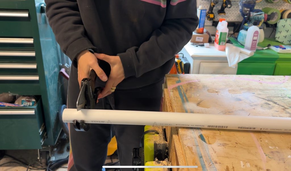 A person uses a pipe cutter to cut a white PVC pipe on a workbench in a workshop, with tools and supplies visible in the background.