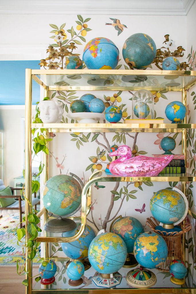 A gold shelving unit displaying a collection of globes, a pink sequined duck, and small sculptures. A leafy plant trails from the top shelf. The wall behind features a colorful, nature-themed mural with birds and lemons.