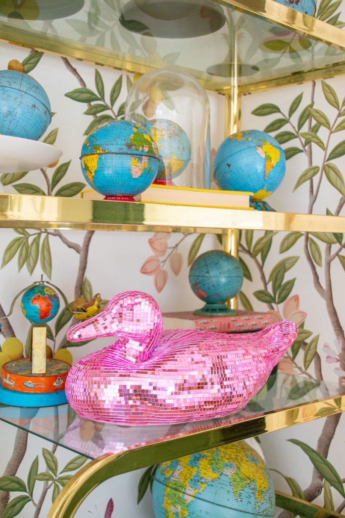 A corner shelf holds multiple decorative globes ranging in size and a shiny pink disco ball duck. The background features wallpaper with illustrated tree branches and leaves. The shelves are accented with gold trim.