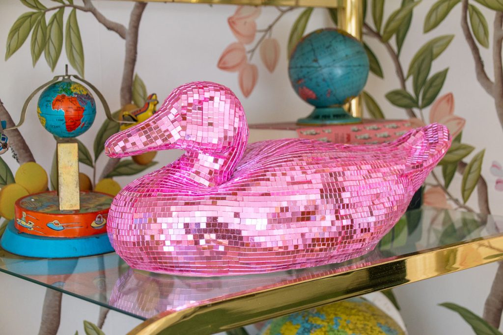 A shiny pink disco mosaic duck figurine sits on a glass shelf. Surrounding it are colorful globes and a tin with illustrated children. The background features a wall with a leafy design, adding a playful and vibrant atmosphere.