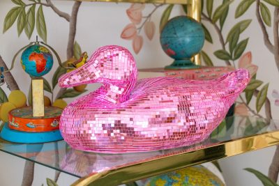 A shiny pink disco mosaic duck figurine sits on a glass shelf. Surrounding it are colorful globes and a tin with illustrated children. The background features a wall with a leafy design, adding a playful and vibrant atmosphere.