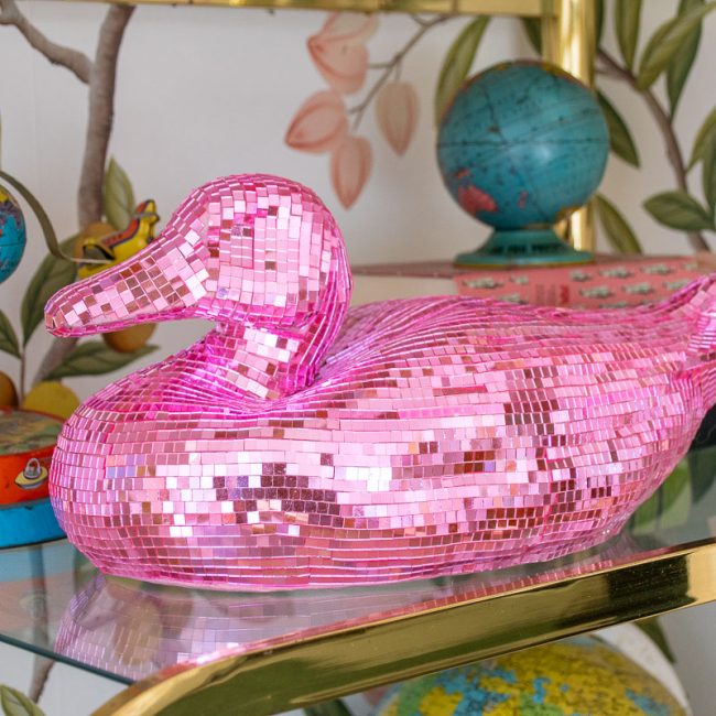 A shiny pink disco mosaic duck figurine sits on a glass shelf. Surrounding it are colorful globes and a tin with illustrated children. The background features a wall with a leafy design, adding a playful and vibrant atmosphere.