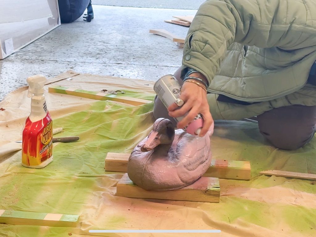 Person kneeling on the floor, spray painting a duck decoy pink. The decoy rests on wooden blocks atop a green sheet. A bottle of cleaning solution is nearby. The person wears a light green jacket inside a garage or workshop.