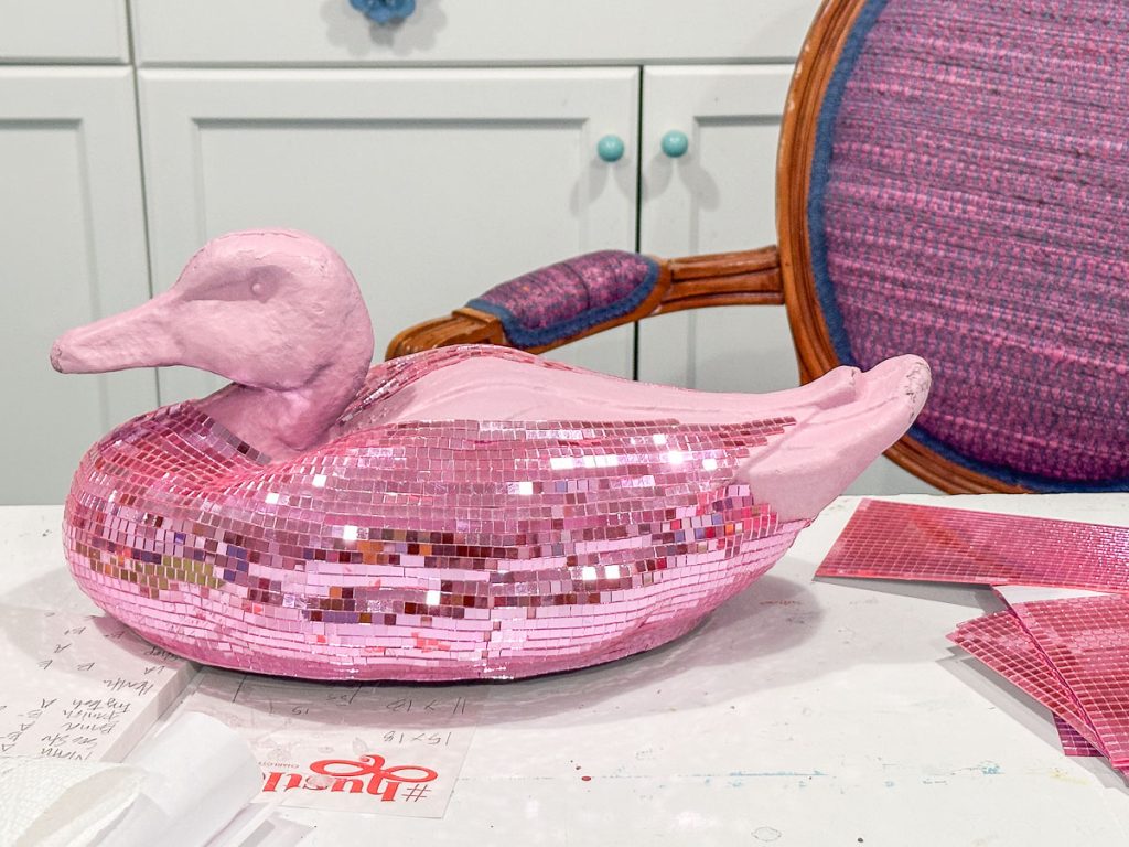 A decorative duck sculpture covered in pink sequins sits on a table. Behind it, a chair with a pink fabric backrest and a white cabinet with turquoise knobs are visible. The table is scattered with papers and pink sequin sheets.