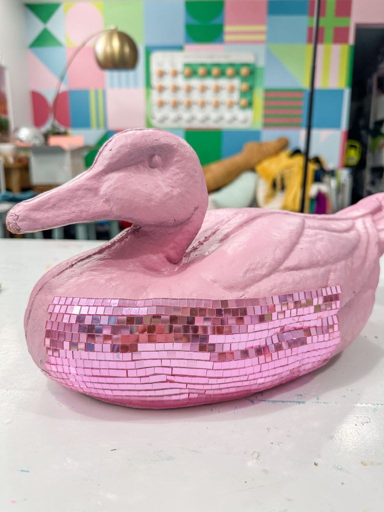 A pink duck-shaped sculpture with a strip of shiny pink mosaic tiles around its body sits on a table. In the background, a room decorated with abstract colorful wall panels, a lamp, and various art supplies is visible.