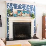 A cozy family room showcases a fireplace with a blue patterned wall above it. Plants and framed artwork adorn the mantel. A colorful rug, green ottoman, and peach couch add vibrant touches to this inviting space.