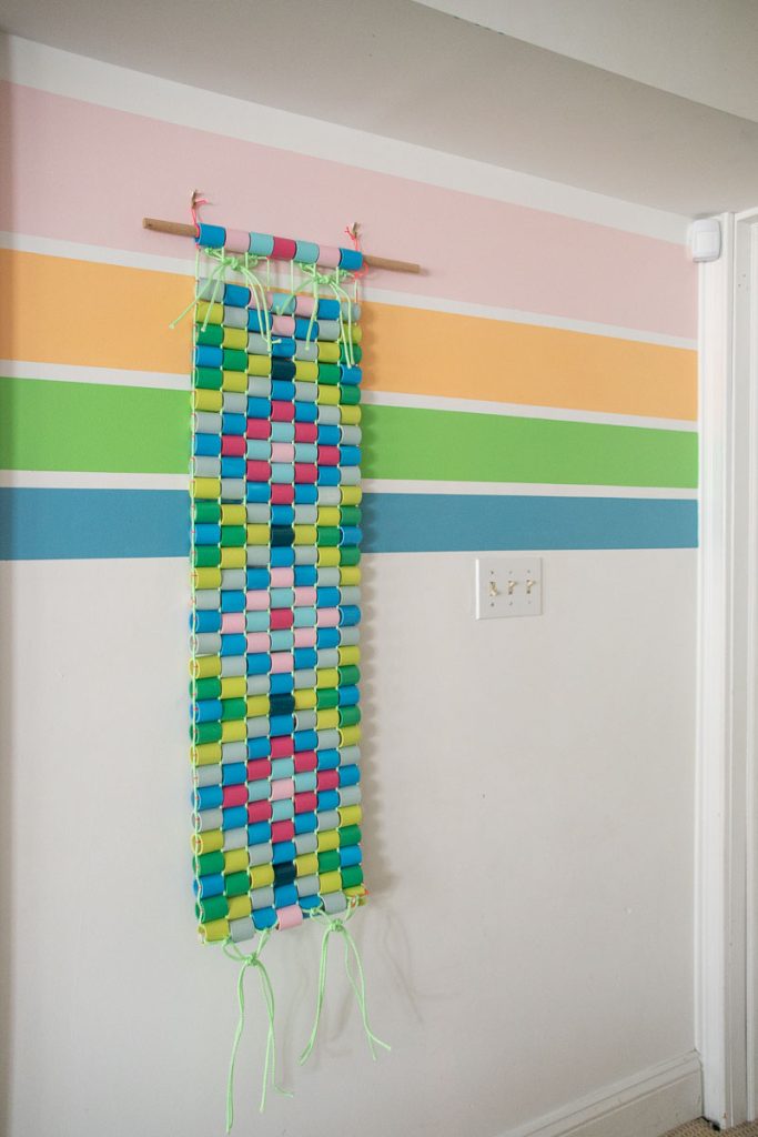 A colorful paper chain wall hanging in blue, green, pink, and yellow hangs from a wooden dowel on a wall with pastel horizontal stripes. Loose strings dangle from the bottom of the hanging.