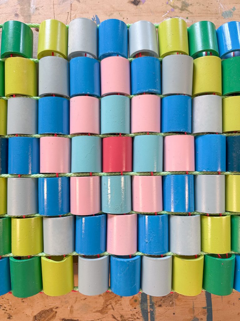Colorful paper chains are arranged in neat horizontal rows, featuring alternating links of blue, green, pink, light gray, and one red link in the center, laying on a wooden surface with visible paint marks.