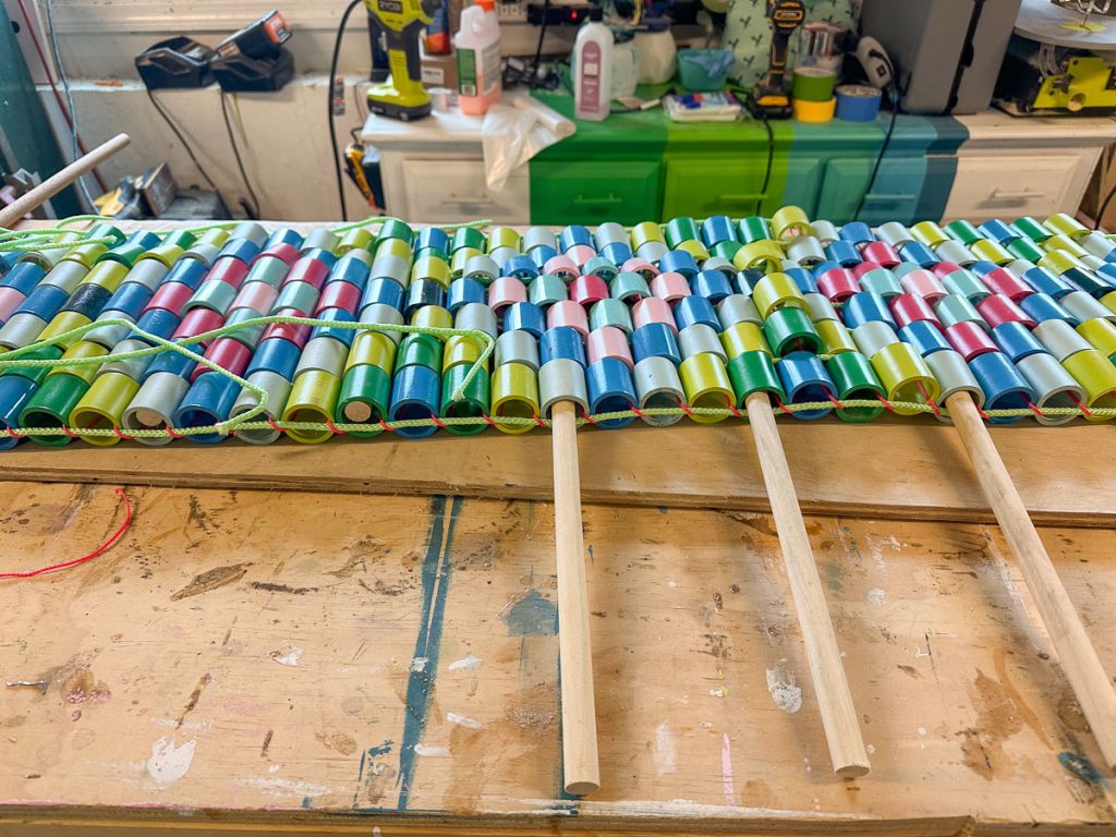 A colorful craft project with rows of plastic spools in blue, green, pink, and white threaded together on a wooden table, with wooden dowels attached underneath. Tools and supplies are visible in the background.