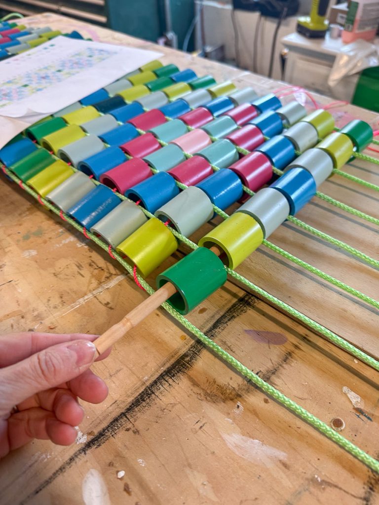 A hand weaves colorful plastic loops onto parallel green strings using a wooden stick, creating a pattern on a wooden tabletop. Craft supplies and a colorful sheet are visible in the background.