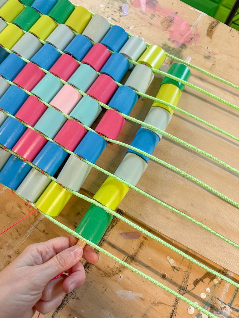 A hand weaving neon green rope through rows of colorful oval beads, arranged in a pattern on a wooden surface. The beads are blue, green, gray, yellow, pink, and red.