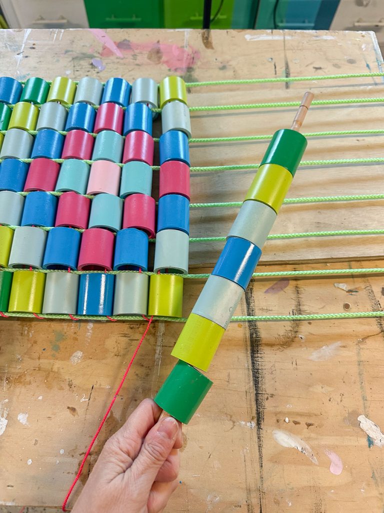 A hand holds a rod with colorful plastic cylinders being threaded onto it. Several rows of similar cylinders are already strung on green cords on a wooden table. The colors include blue, green, pink, and yellow.