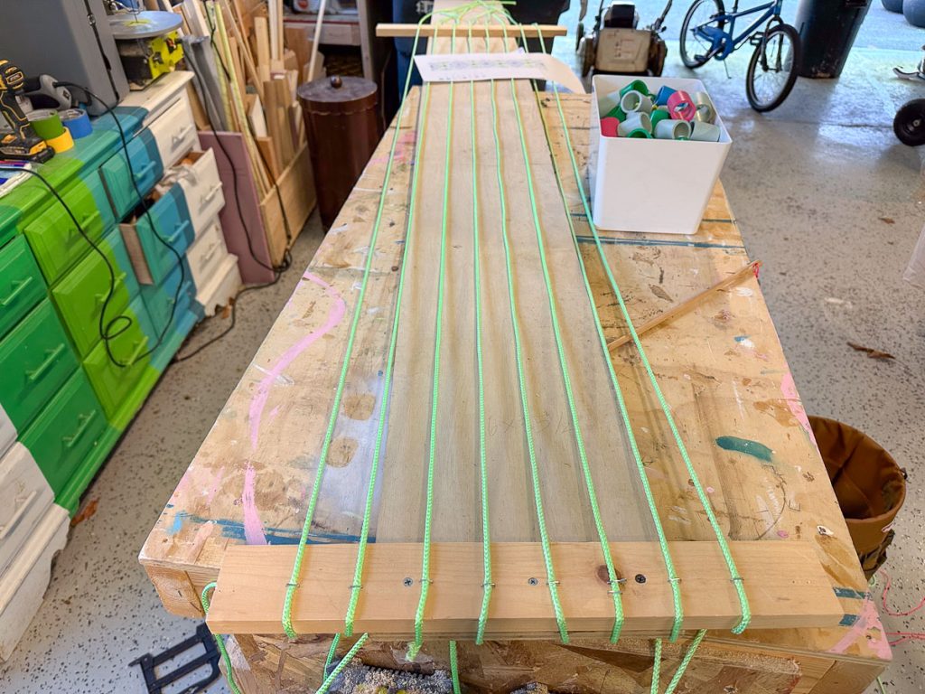 A wooden board on a workbench has green string stretched and tied across it in parallel lines. Workshop tools and materials are visible in the background.