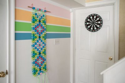 A colorful paper chain decoration hangs on a pastel-striped wall next to a white door with a dartboard mounted on it. The scene is bright and playful, with a staircase visible in the bottom right corner.