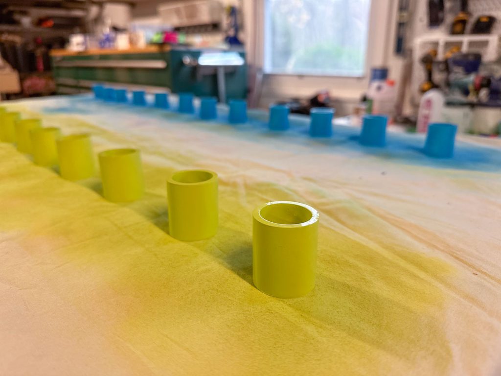 Several small cylindrical objects, some lime green and others blue, are arranged in rows on a surface with a gradient of yellow and blue colors in a workshop or studio setting.