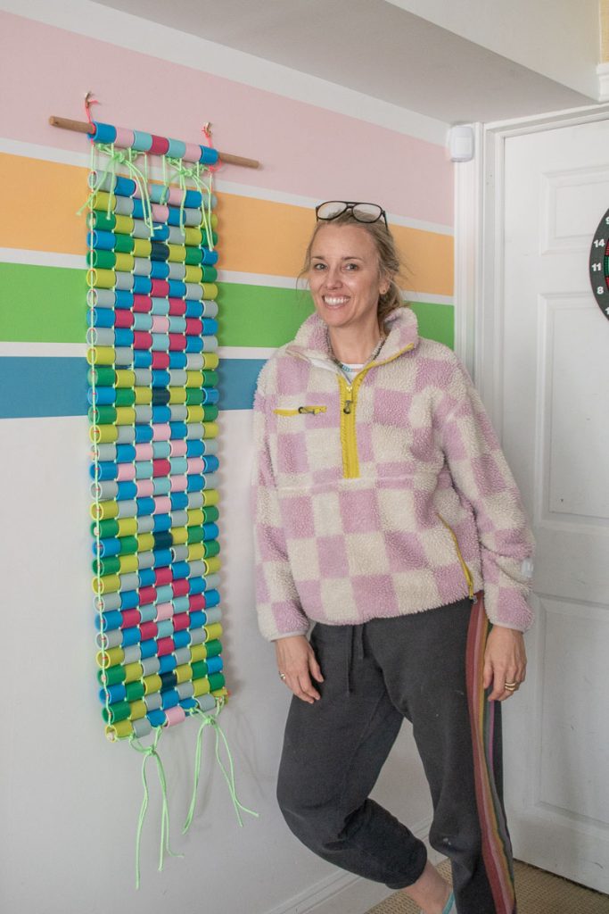 A smiling woman stands indoors beside a colorful paper chain wall hanging on a wooden dowel. The wall behind her has horizontal pastel stripes. She is wearing a checkered fleece jacket and dark pants.