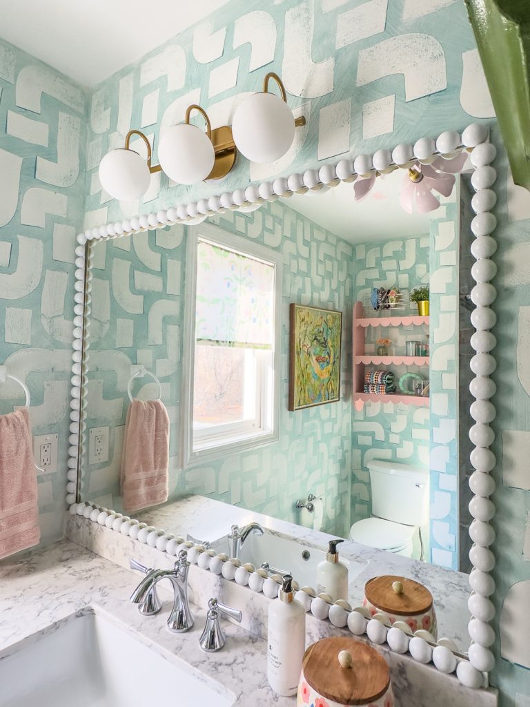 A bathroom with light blue geometric-patterned walls, a large mirror with a white beaded frame, and a marble countertop. There are pink towels, a wall-mounted light fixture with three bulbs, and a shelf with decorative items. A window lets in natural light.