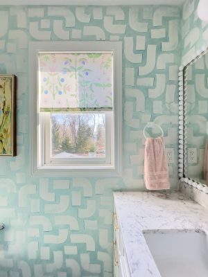 Bathroom with patterned mint-green walls, a window with floral-patterned blinds, and a marble countertop. A pink towel hangs by a circular holder. A large mirror with a white beaded frame reflects soft light.