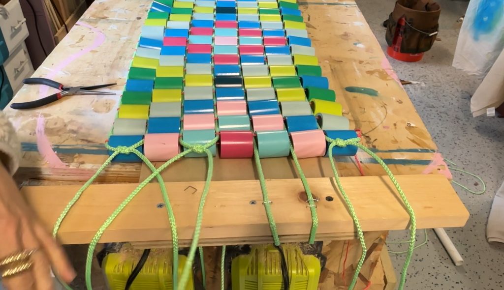 Colorful plastic tiles are woven together with green ropes on a wooden frame, resting on a workbench. Hands are seen at the sides, suggesting an ongoing crafting or weaving project.